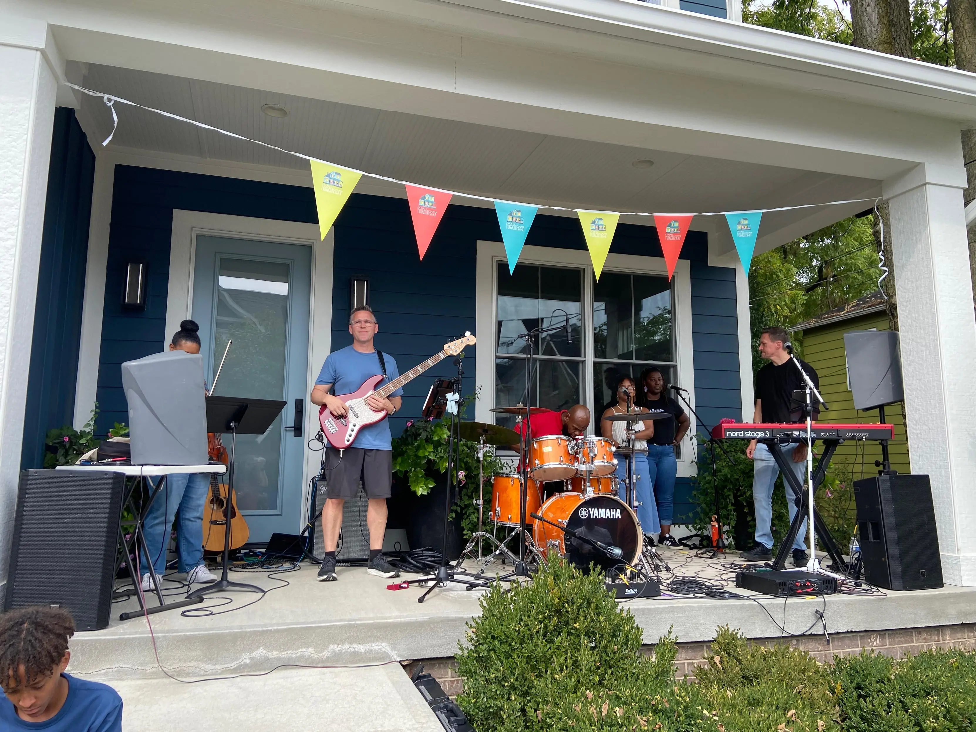 A small band standing on a porch. From left to right, there is a violinist,
bassist, drummer, two vocalists, and a keyboardist/vocalist.
