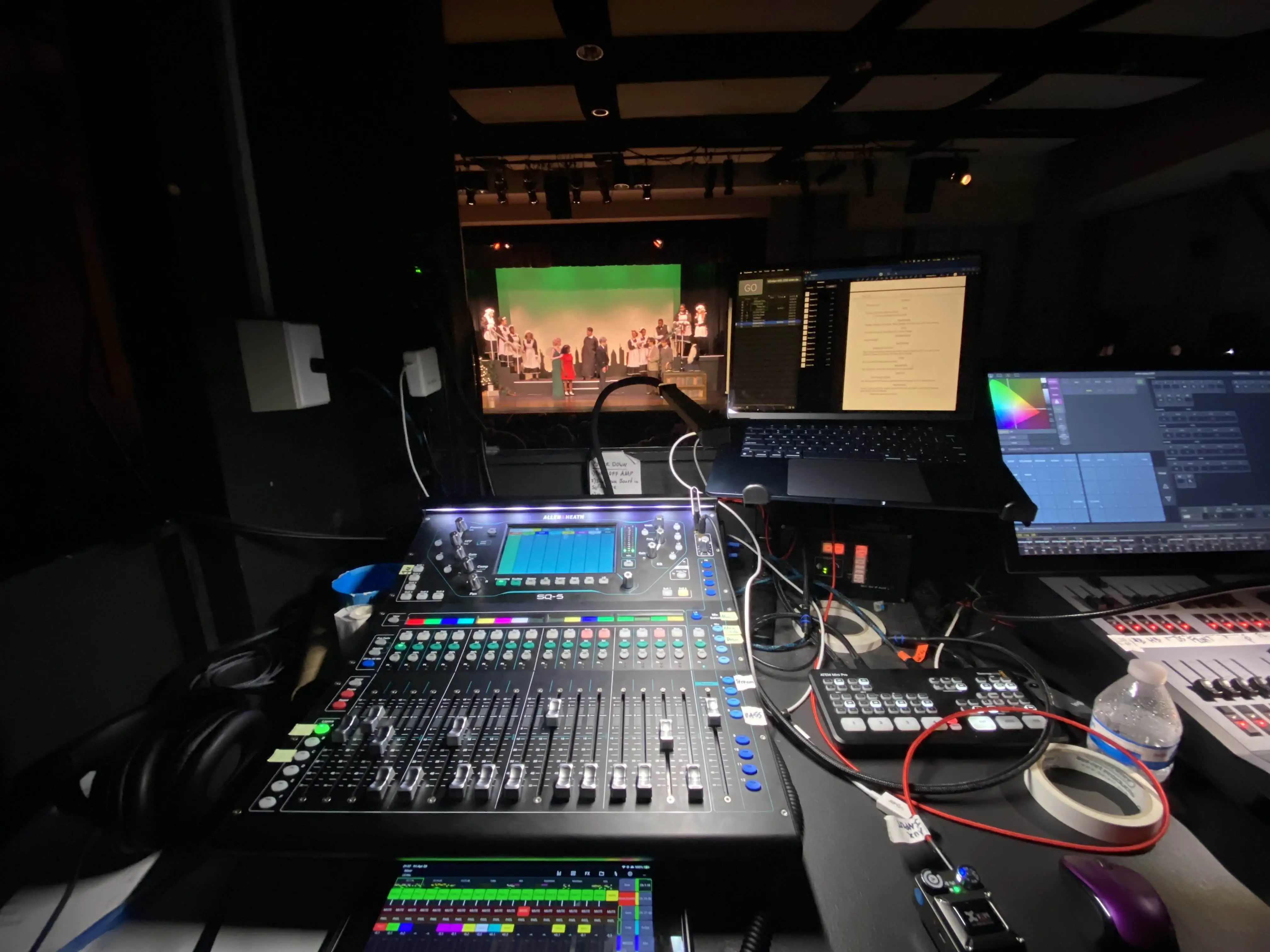A view of a mixer and laptop on a desk, with a stage in the background.