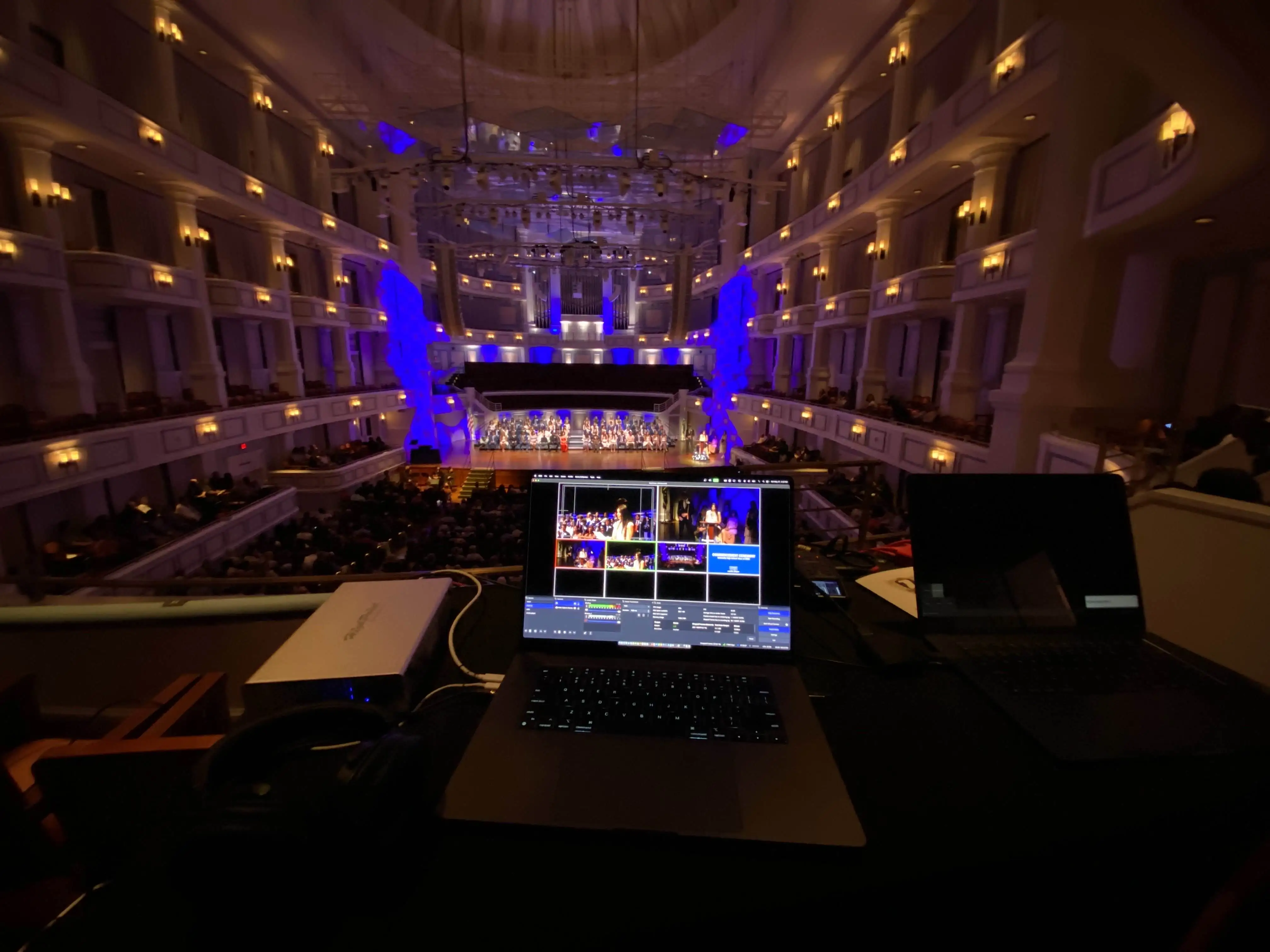 A view of a desk with two laptops and other miscellaneous video eqipment,
overlooking a large hall.
